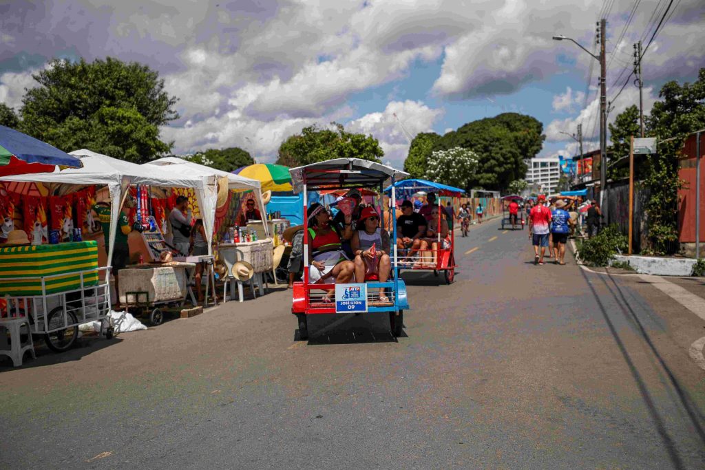 Visitantes passeiam de triciclo durante o Festival de Parintins 2024, com vista para o rio Amazonas