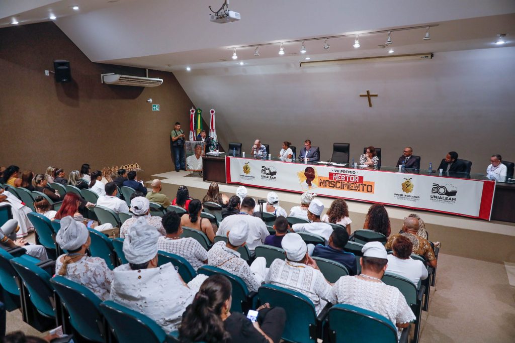 Prêmio Nestor Nascimento Aleam celebra luta pela igualdade racial no Amazonas Pessoas sentadas em um auditório, de costas para a câmera, com várias delas usando roupas e adereços brancos que remetem a religiões de matriz africana. Ao fundo, uma mesa com autoridades e um banner com o título 'VII Prêmio Nestor Nascimento' e logos de instituições.
